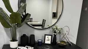 A modern desk with a large mirror, a potted plant, candles, a framed picture, and a small table with a bowl and utensils.