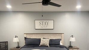 A bedroom with a bed, two bedside tables, a ceiling fan, a wall-mounted sign, and a patterned rug.