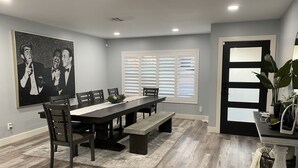 A dining area with a long table, chairs, a bench, a painting, a window with blinds, and a door with frosted glass panels.