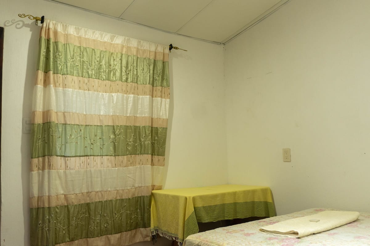 Ein kleines, schlichtes Zimmer mit einem Bett, einem Tisch und einem Vorhang.