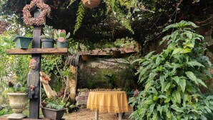 Ein rustikaler Garten im Freien mit einem Holztisch, bedeckt mit einer gelben Tischdecke, umgeben von verschiedenen Topfpflanzen und einer dekorativen Holzfigur.