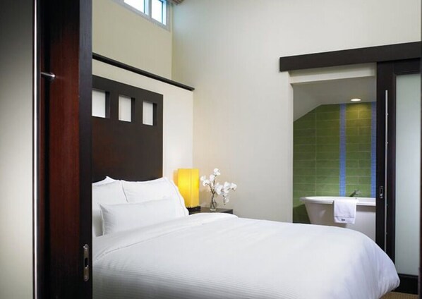 Habitación de hotel con cama, mesita de noche con lámpara, un jarrón con flores y vistas al baño.