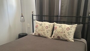 Cama bem arrumada com almofadas florais, mesa de cabeceira com abajur e janela com cortinas.
