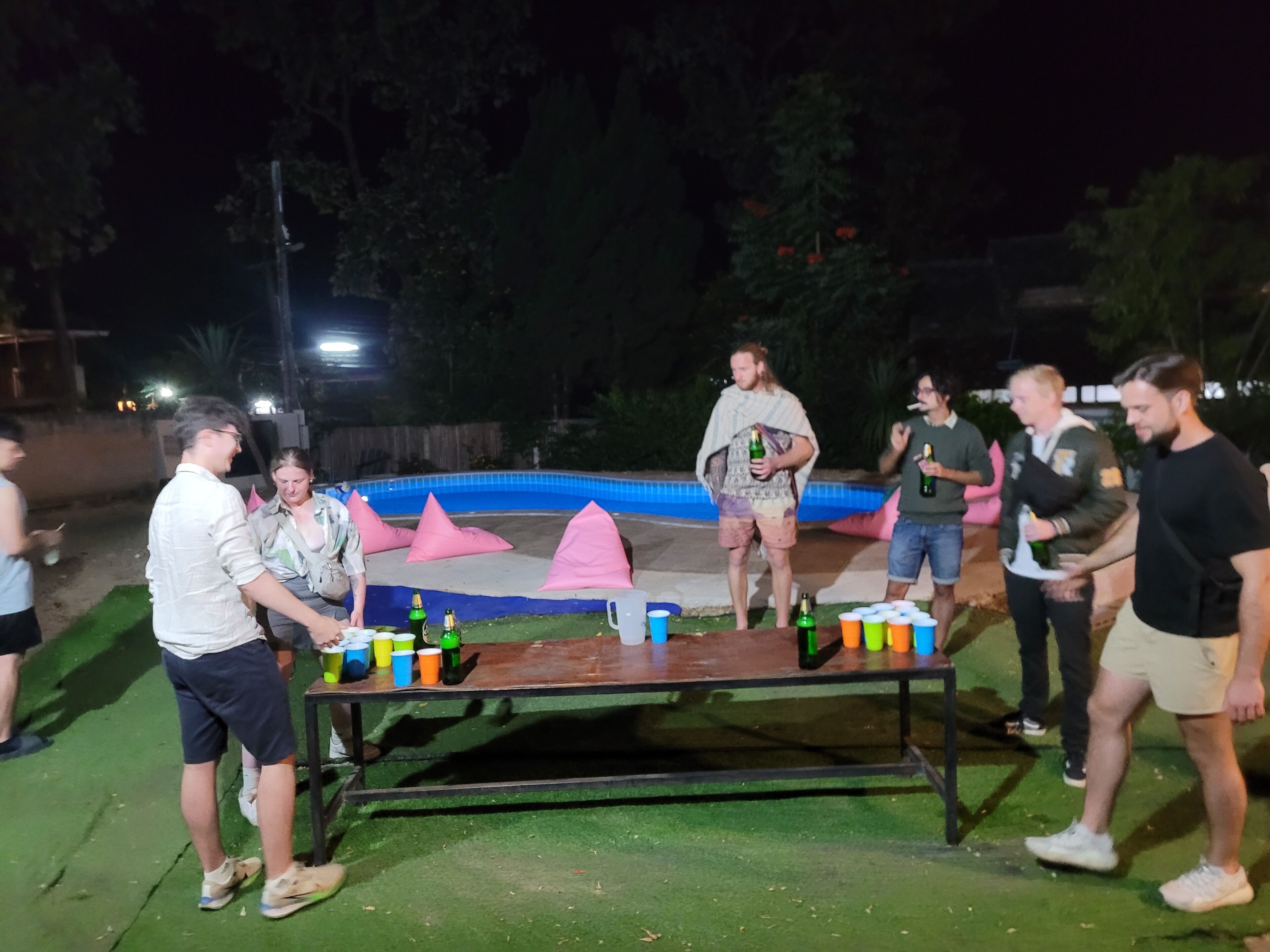 A group of people around a table with bottles and cups, near a pool with pink cushions.