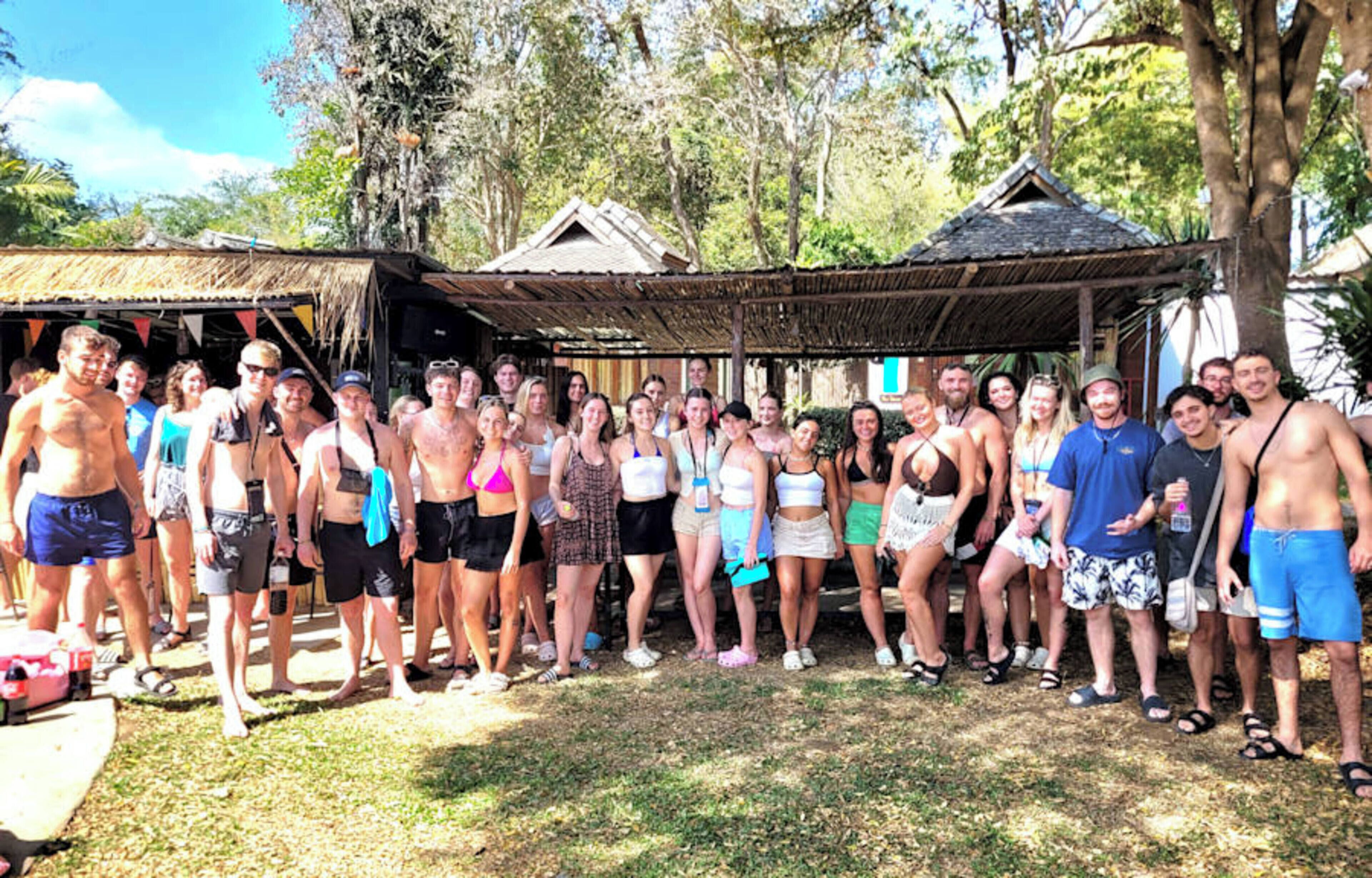 A group of people standing outdoors in a tropical setting with thatched-roof structures and lush greenery.