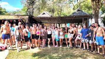 A group of people standing outdoors in a tropical setting with thatched-roof structures and lush greenery.