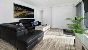 A modern living room with a black leather sofa, a glass coffee table, and a large painting of a coastal landscape.