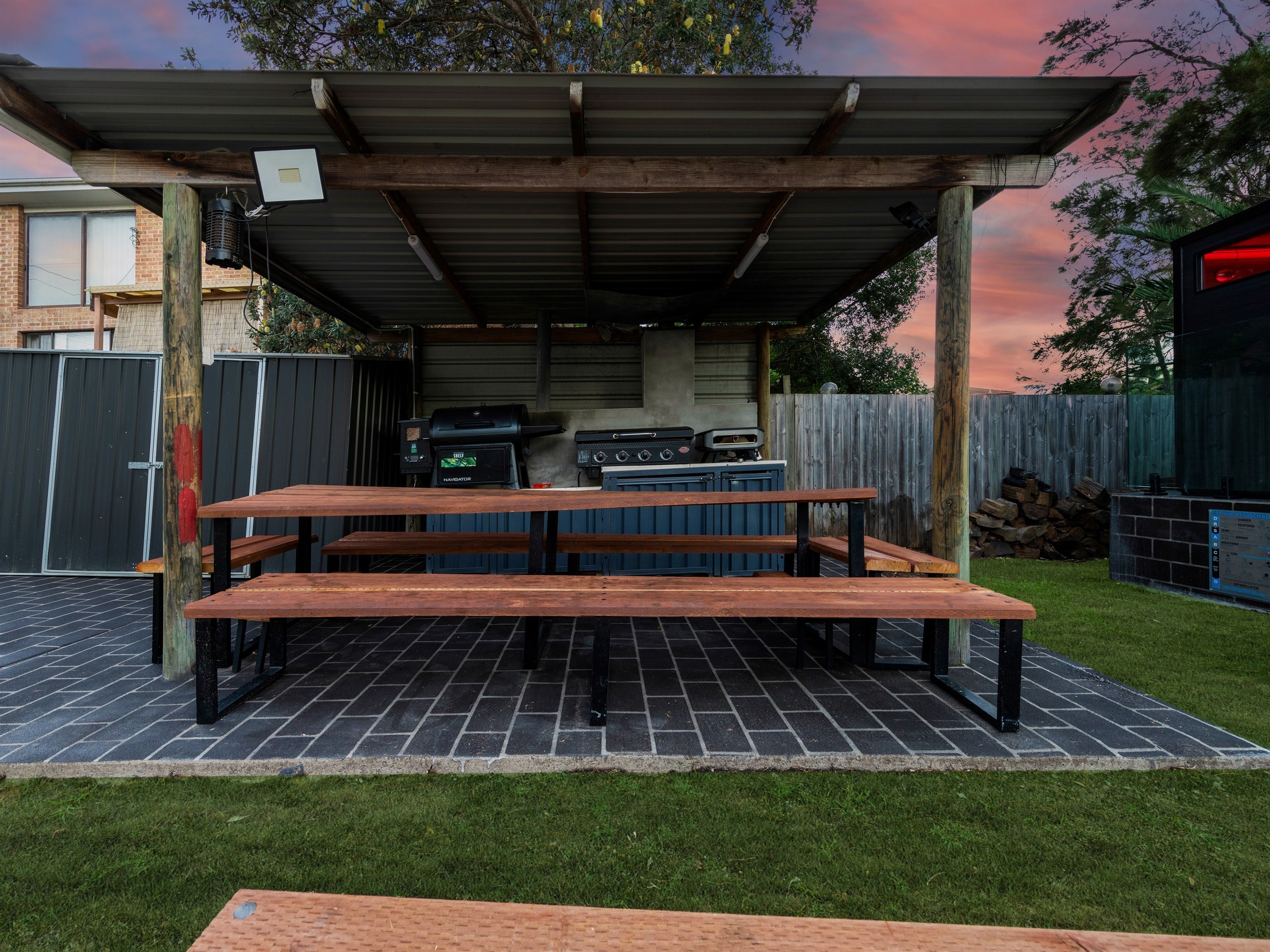 An outdoor barbecue area with a covered grill station, picnic tables, and a brick patio.