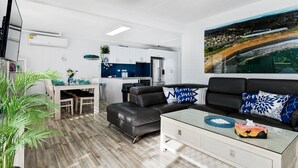A modern living room with a black leather sofa, a glass coffee table, and a dining area with a white table and chairs. A kitchen is visible in the background with white cabinets and blue backsplash. - SKYES BEACH HOUSE | spas | sauna | arcade machines (Malua Bay)
