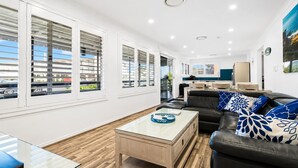 A modern living room with a glass coffee table, black leather sofa, and a view of the street through large windows. - SKYES BEACH HOUSE | spas | sauna | arcade machines (Malua Bay)
