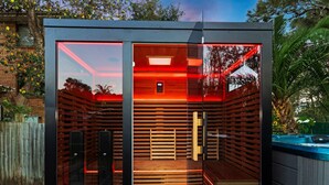 A modern sauna with glass walls, wooden benches, and red lighting. - SKYES BEACH HOUSE | spas | sauna | arcade machines (Malua Bay)