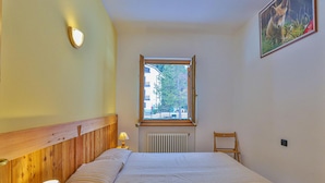 Un dormitorio con cabecera de madera, cama con ropa de cama a rayas, mesita de noche con lámpara, ventana con vistas a edificios y árboles, y un cuadro en la pared.