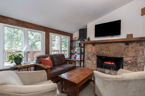 A living room with a stone fireplace, a flat-screen TV, a bookshelf, and a window with a view of trees. - Manitou Lodge 8 Hotel Room by Alpine Lodging Telluride (Telluride)