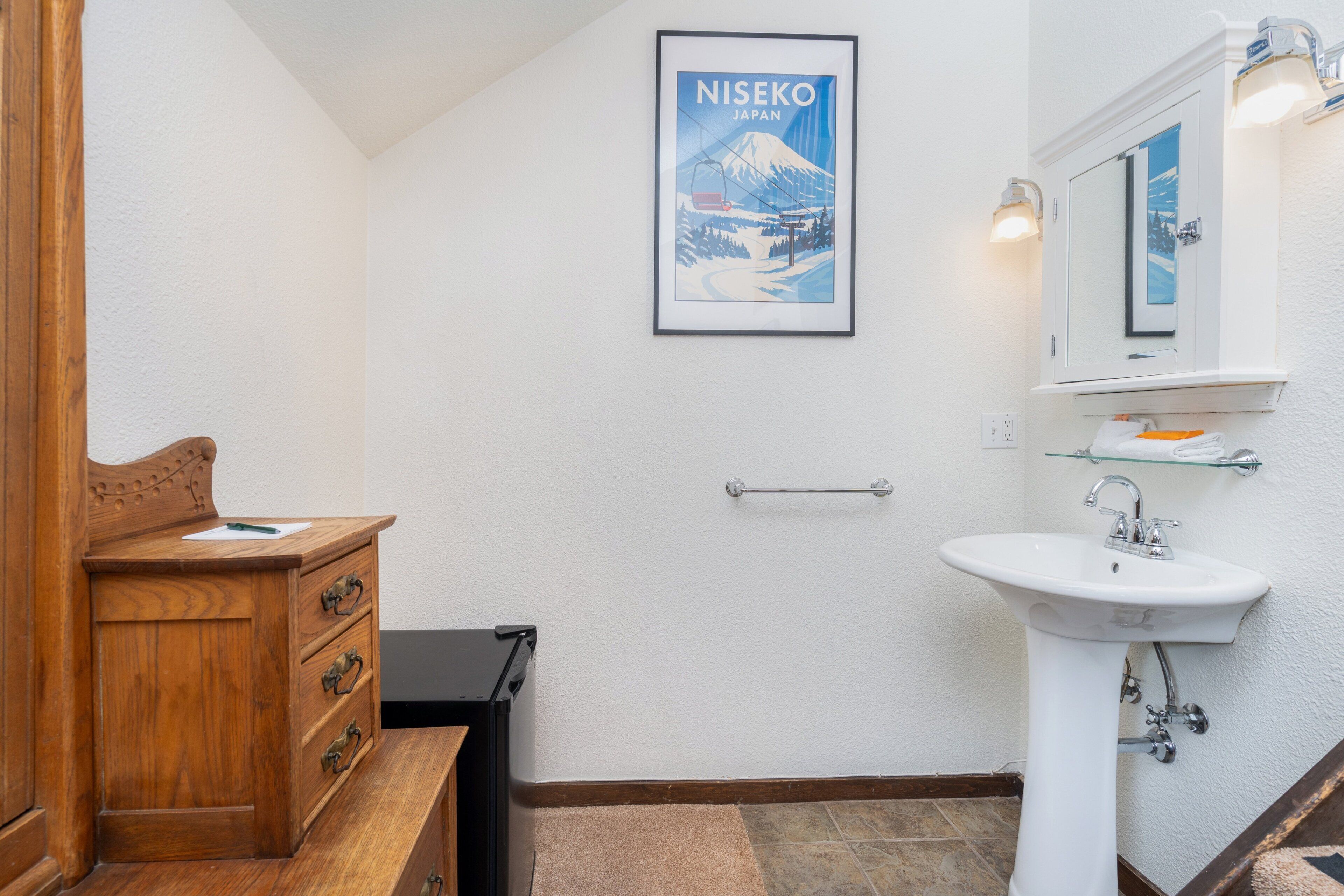 A bathroom with a wooden vanity, a white sink, a framed poster of Niseko, Japan, and a mirror with lights.