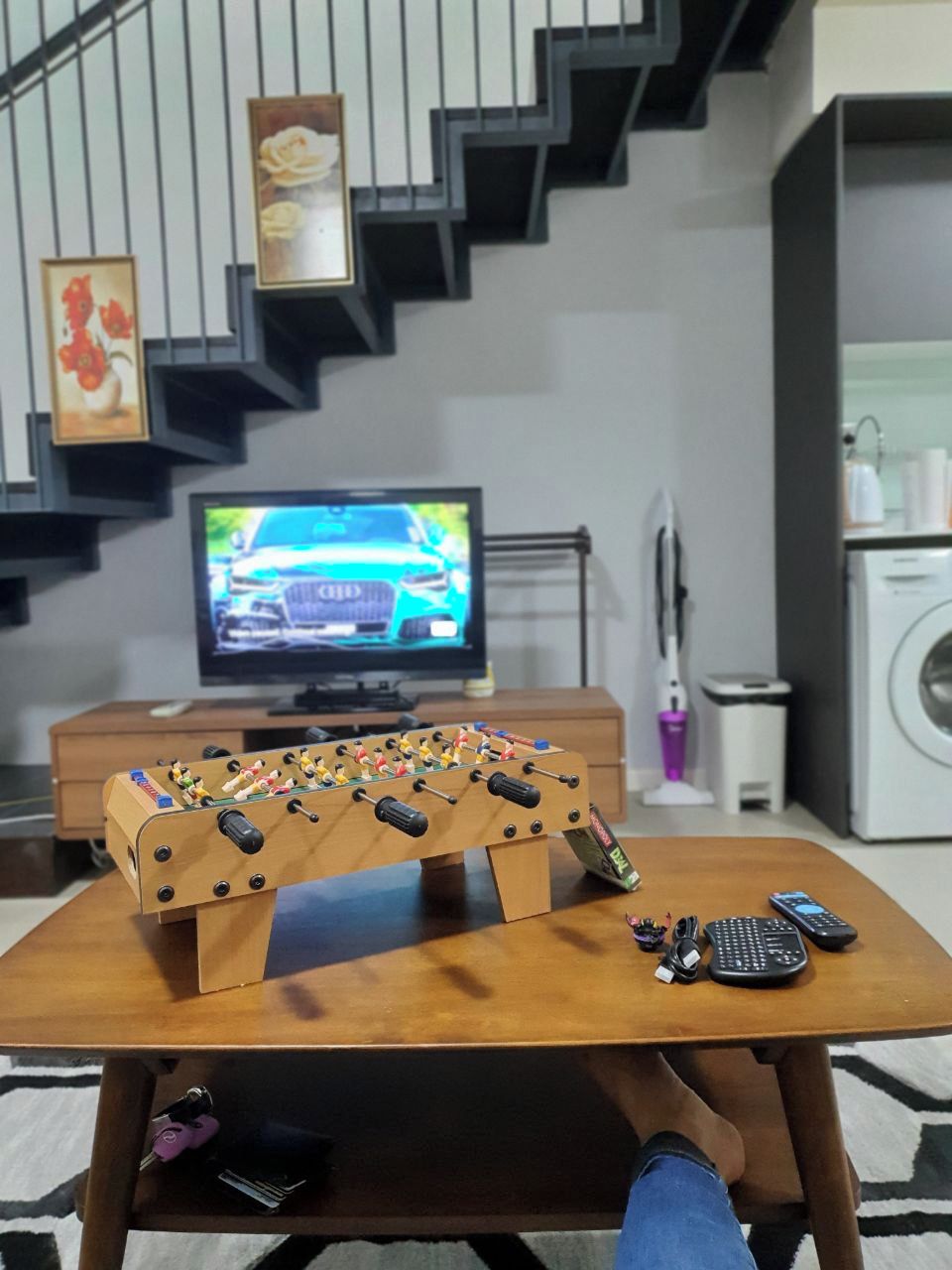 Una sala de estar con una mesa de fútbol de mesa, un televisor mostrando un automóvil y una lavadora.