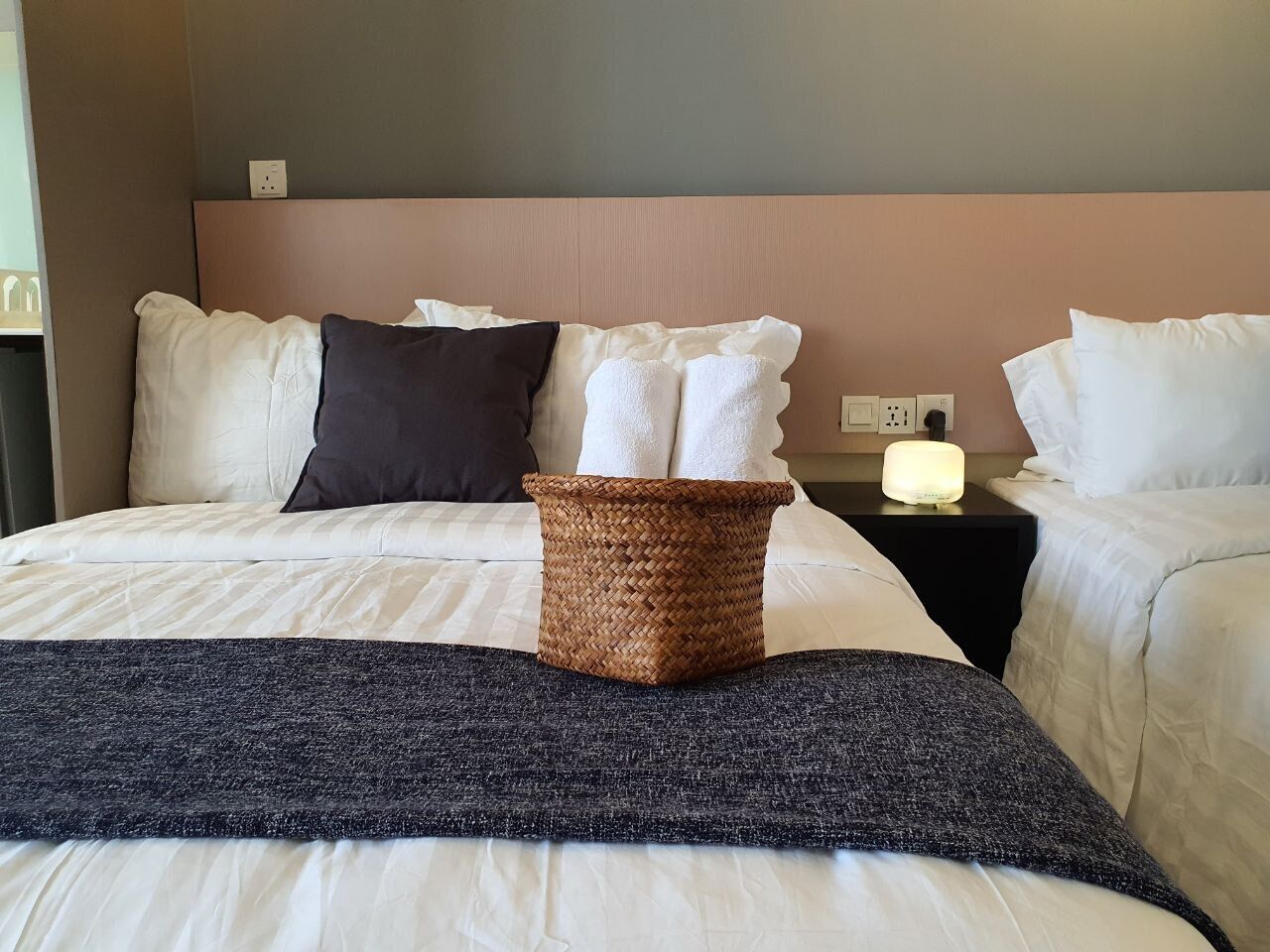 A hotel room with a bed, pillows, a woven basket, and a bedside table with a lamp.