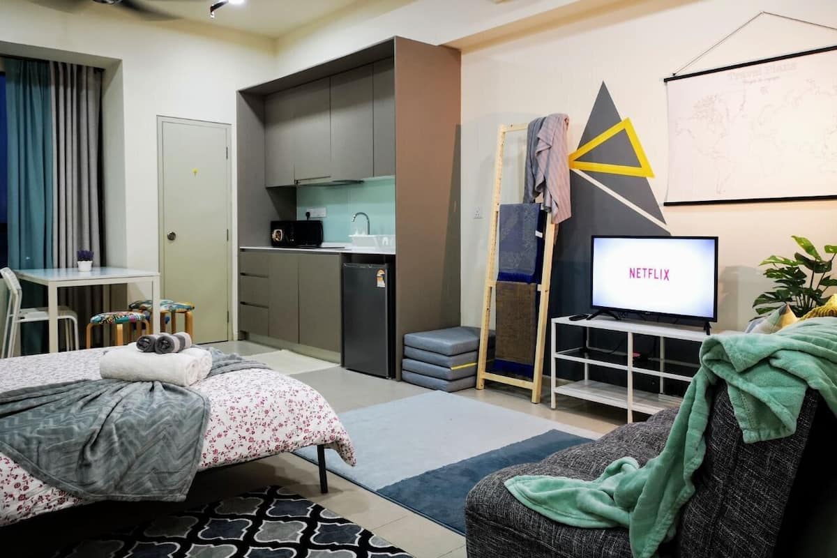 A compact hotel room with a bed, a desk, a kitchenette, and a TV displaying 'NETFLIX'.