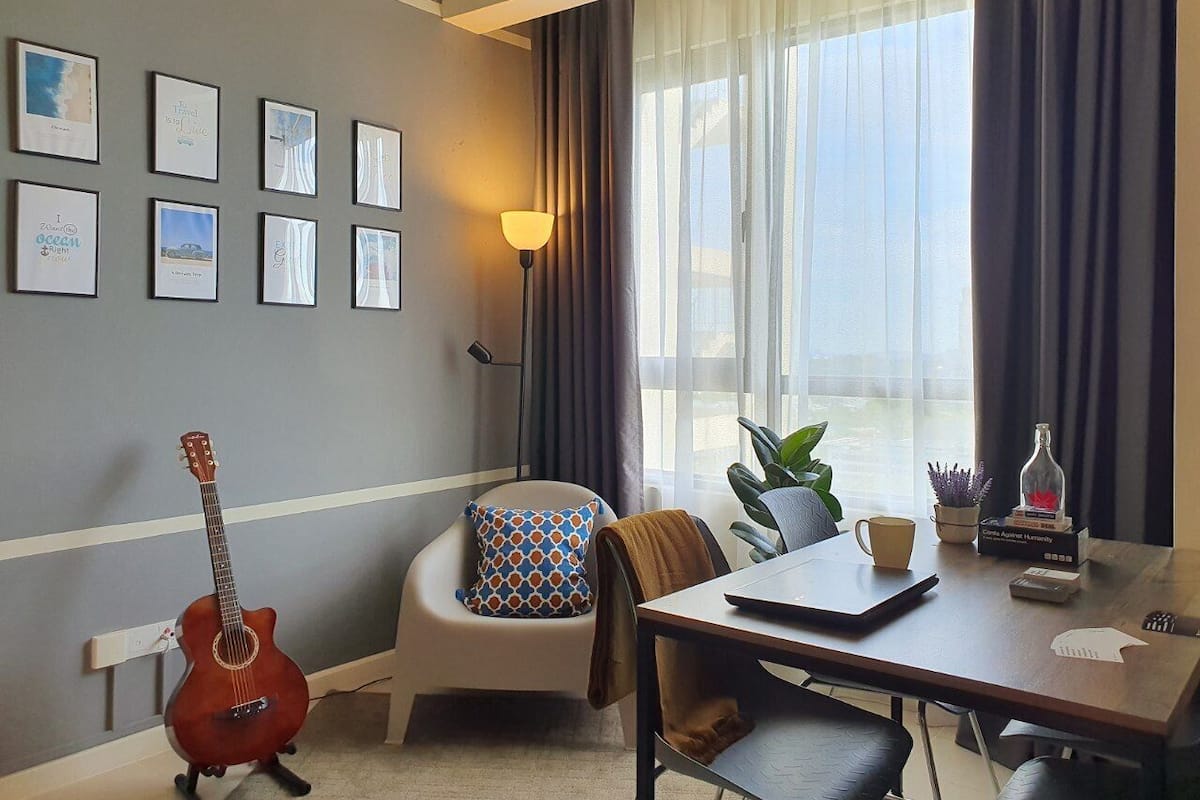 A room with a dining table, a guitar, a chair with a patterned cushion, a lamp, and framed pictures on the wall.