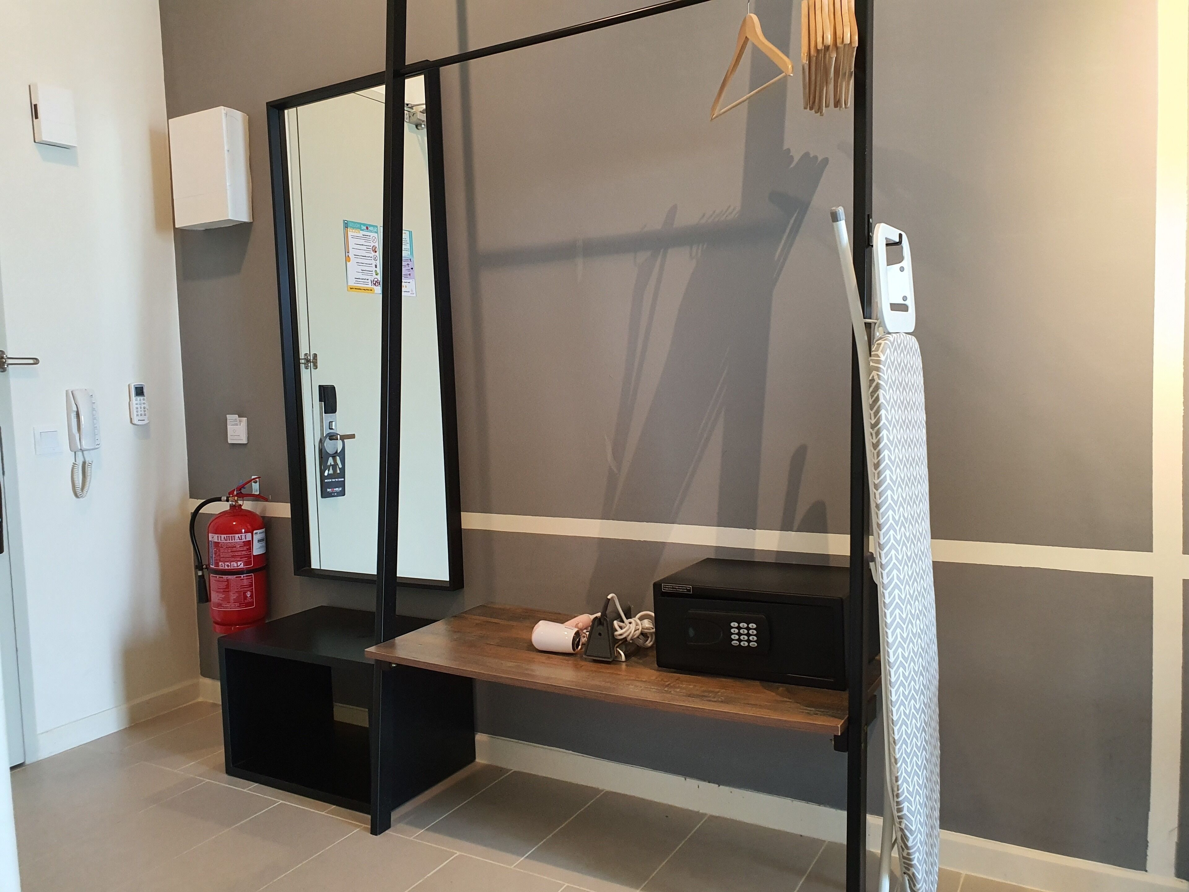 A hotel room with a wooden bench, a black electronic device, a fire extinguisher, and a mirror on the wall.