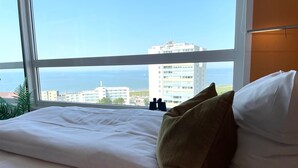 Une chambre d’hôtel avec une grande fenêtre, un lit avec du linge de maison blanc, un oreiller marron et une vue sur la ville et la mer.