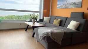 Un salon moderne avec un canapé gris, une table basse en bois et une grande fenêtre donnant sur un paysage verdoyant et un plan d’eau.