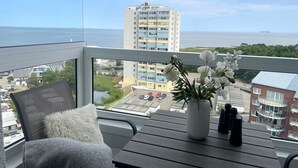 Un balcon donnant sur la mer, une chaise, une table avec un vase de fleurs et un bâtiment en arrière-plan.