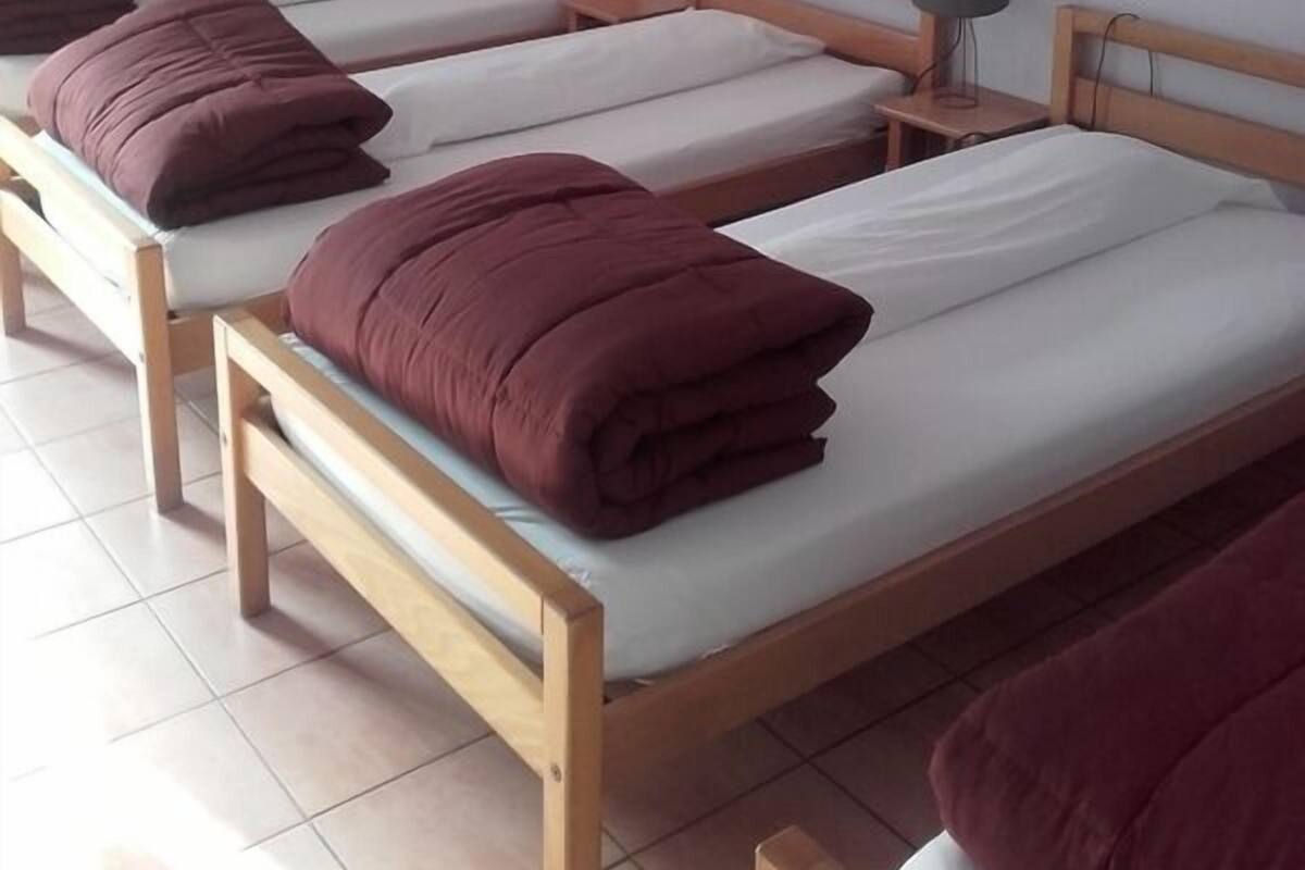 Ein Zimmer mit vier Holzbetten, jedes mit einer weißen Matratze und einer braunen Decke. An einer Seite befindet sich ein kleiner Nachttisch mit einer Lampe.