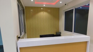 A reception desk with a white top and a wooden panel, a window with blinds, and a wall with vertical lines and a small screen. - Amada Hotel (Lahore)