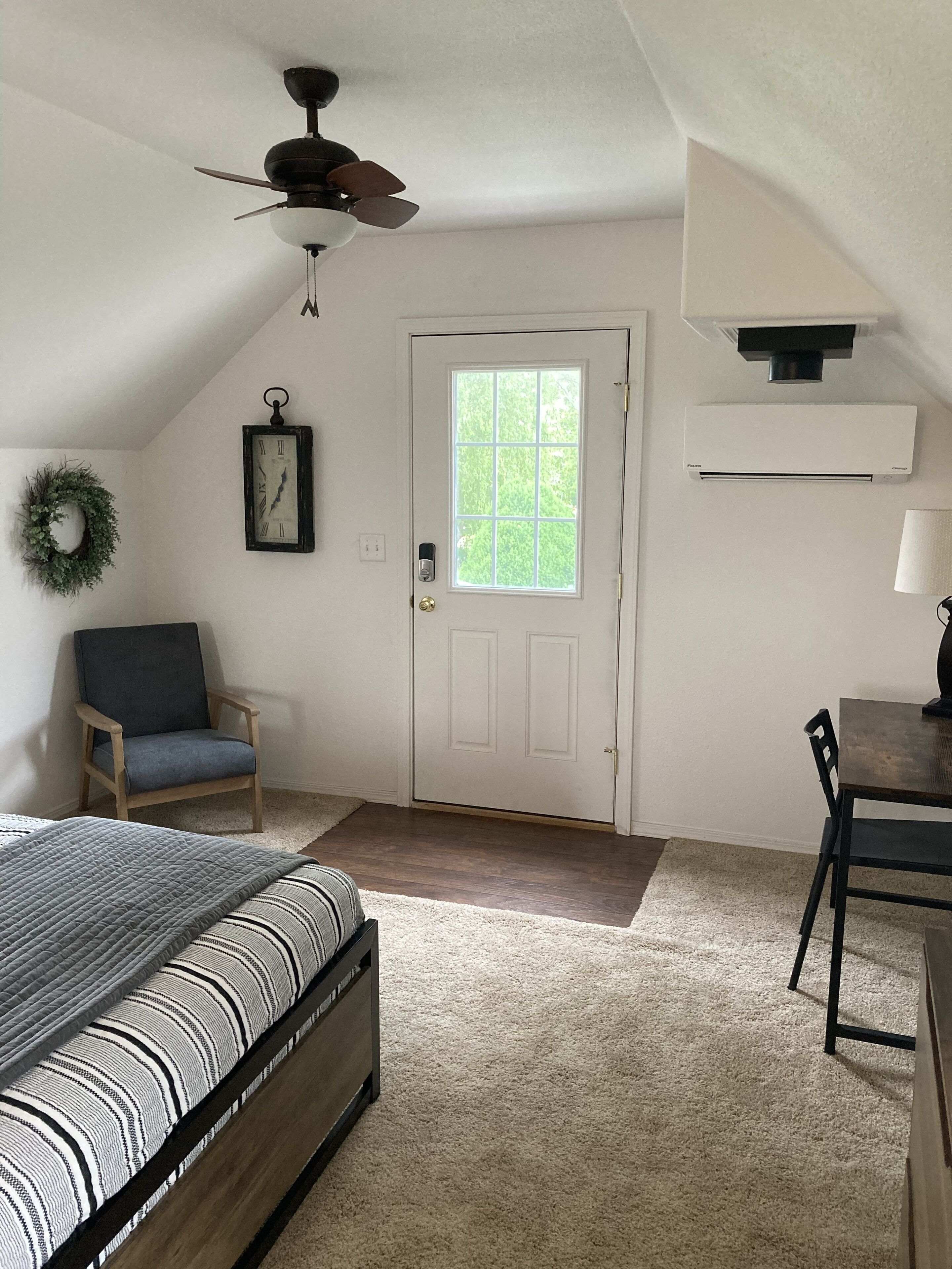 Un dormitorio con cama, una silla, un escritorio, ventilador de techo, puerta con ventana y un reloj en la pared.