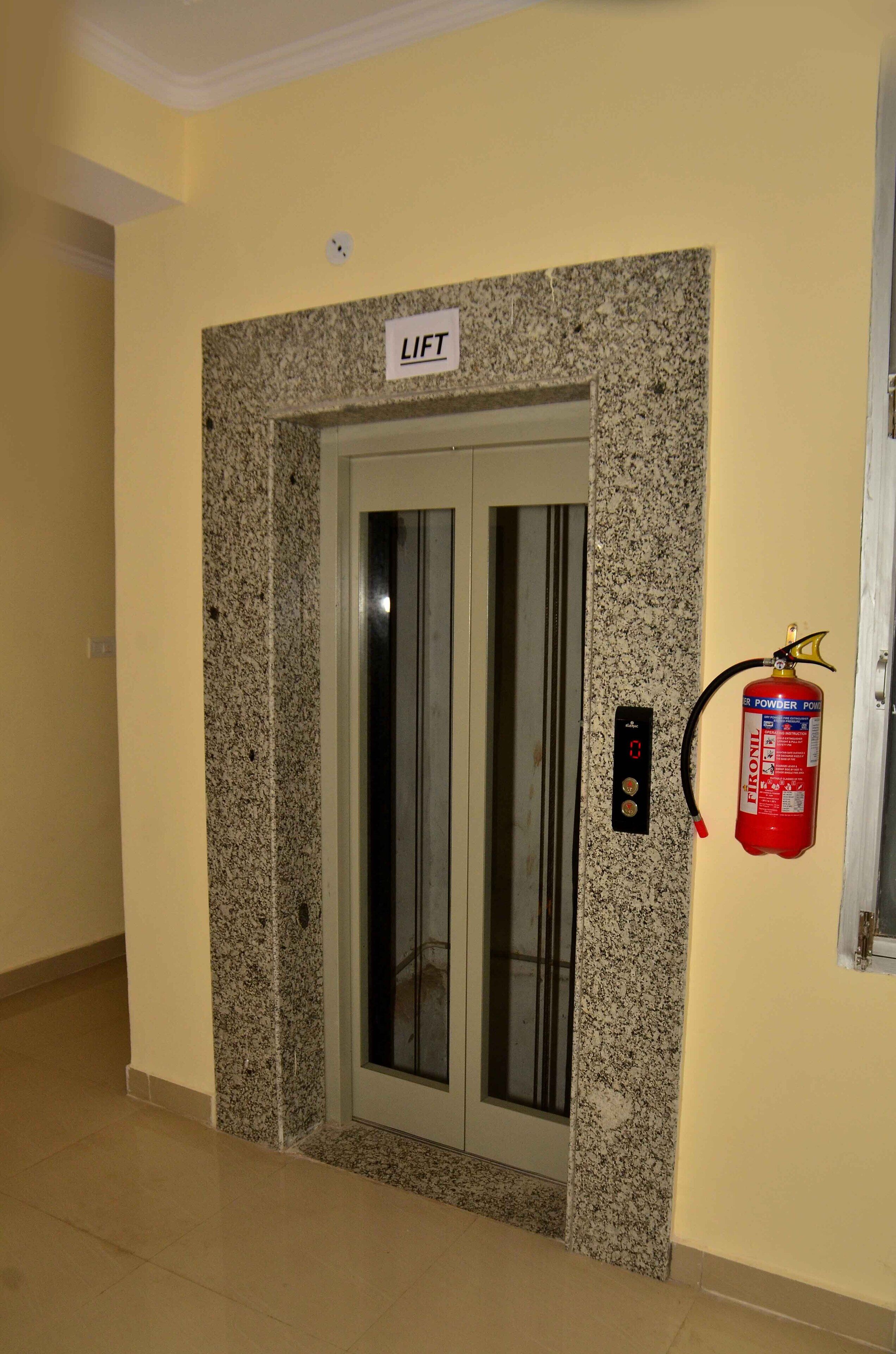 Ein Aufzug mit einem Rahmen aus Granit, einem an der Wand befestigten Feuerlöscher und einem Schild über der Tür mit der Aufschrift 'LIFT'.
