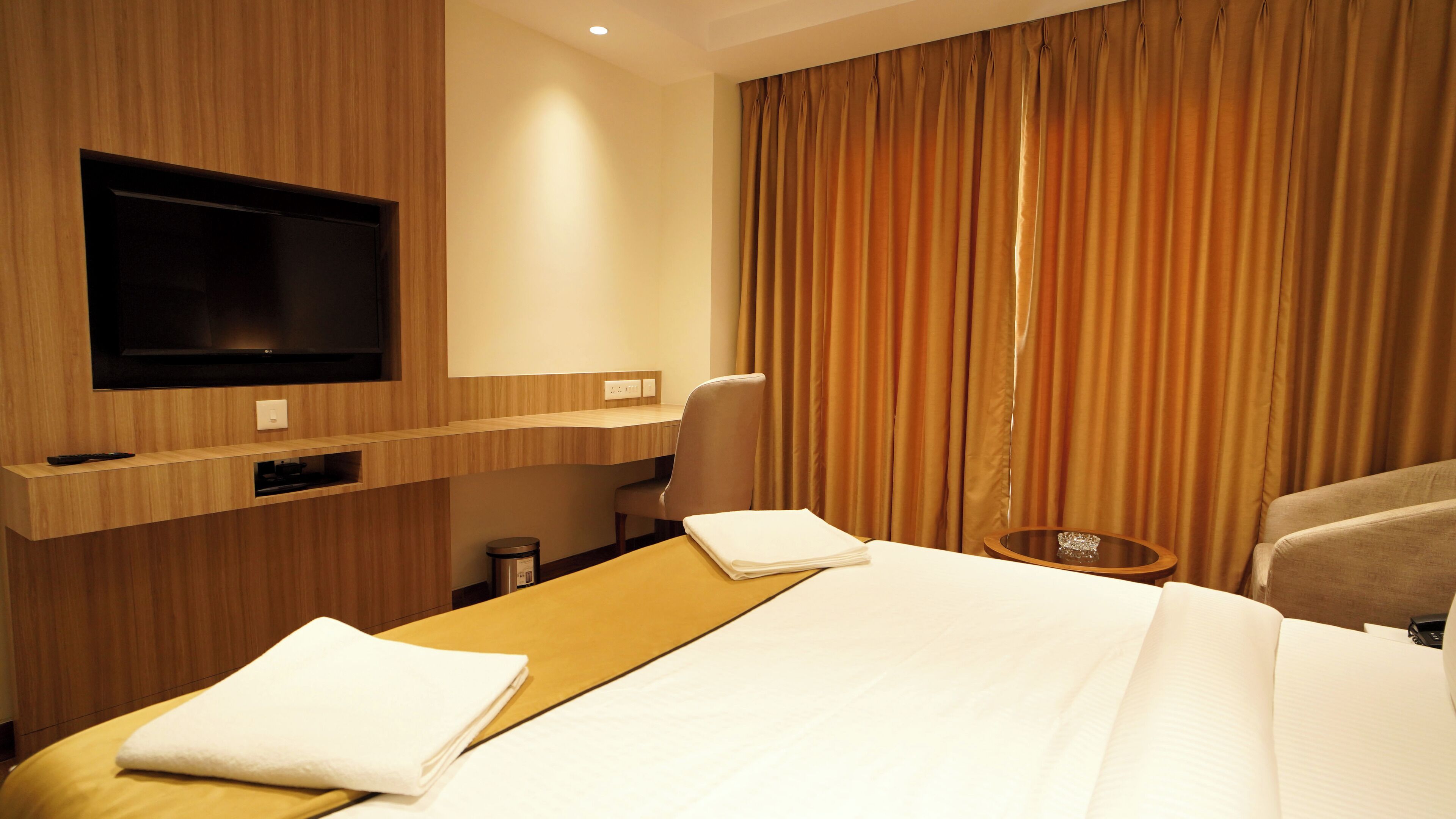 A hotel room with a bed, a TV mounted on the wall, a desk with a chair, and a window with curtains.