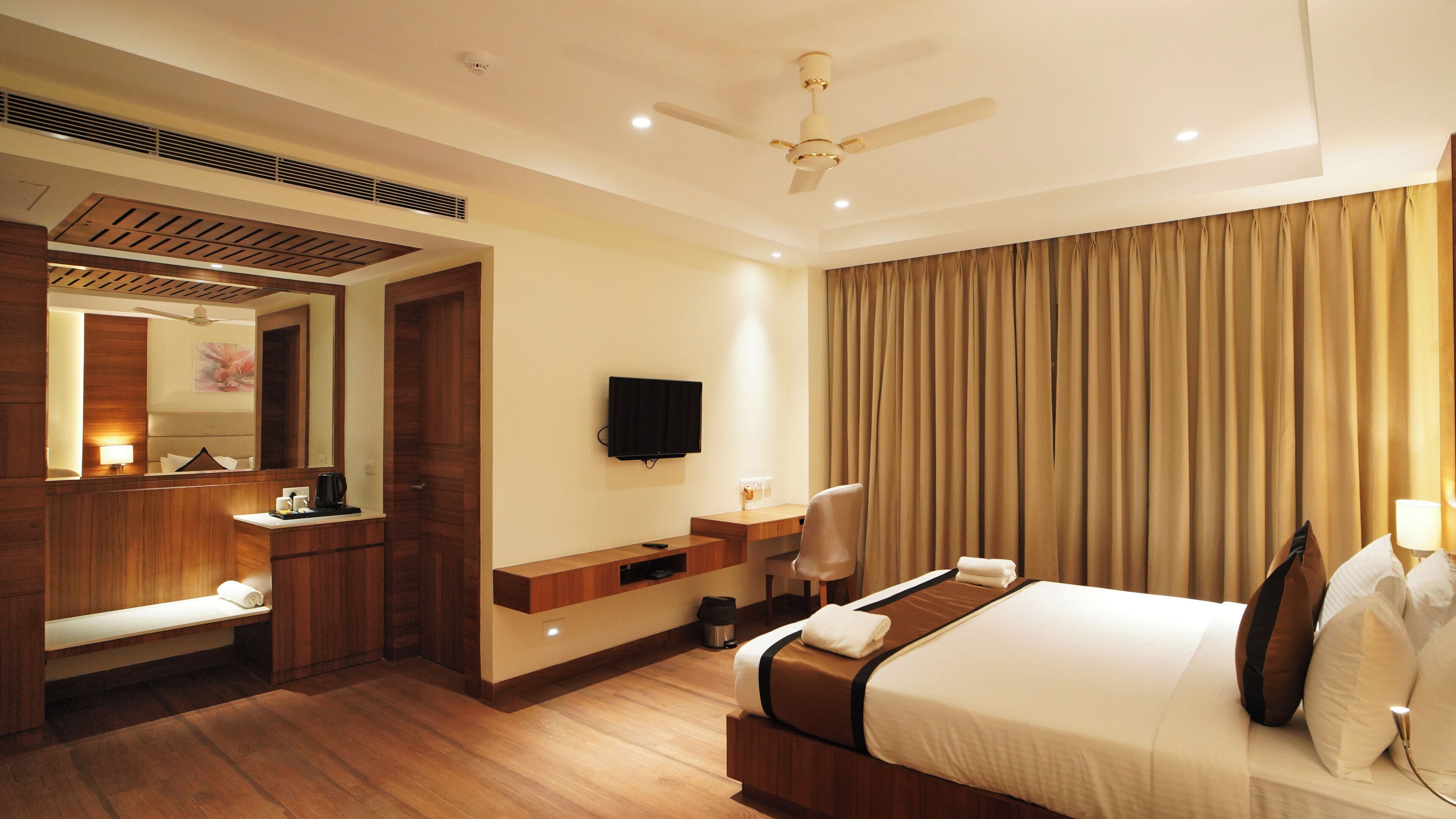 A hotel room with a bed, a desk, a TV, a mirror, and a ceiling fan.