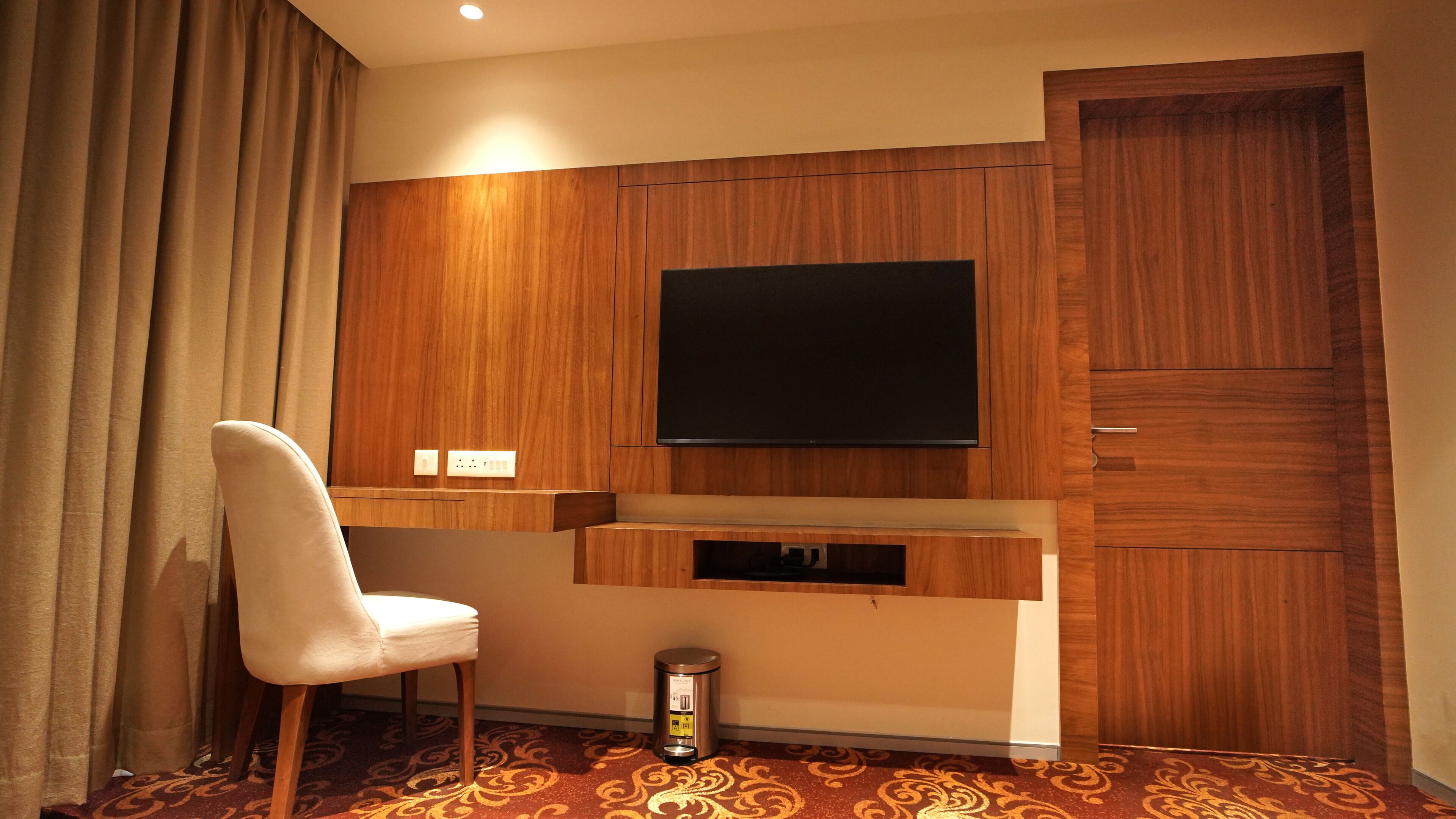 A hotel room with a wooden panel wall, a flat-screen TV mounted above a wooden console, a white chair, and a trash bin.