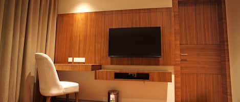 Ein Hotelzimmer mit einer Wandpaneele aus Holz, einem flachbildfernseher über einer Holzkonsole, einem weißen Stuhl und einem Mülleimer.