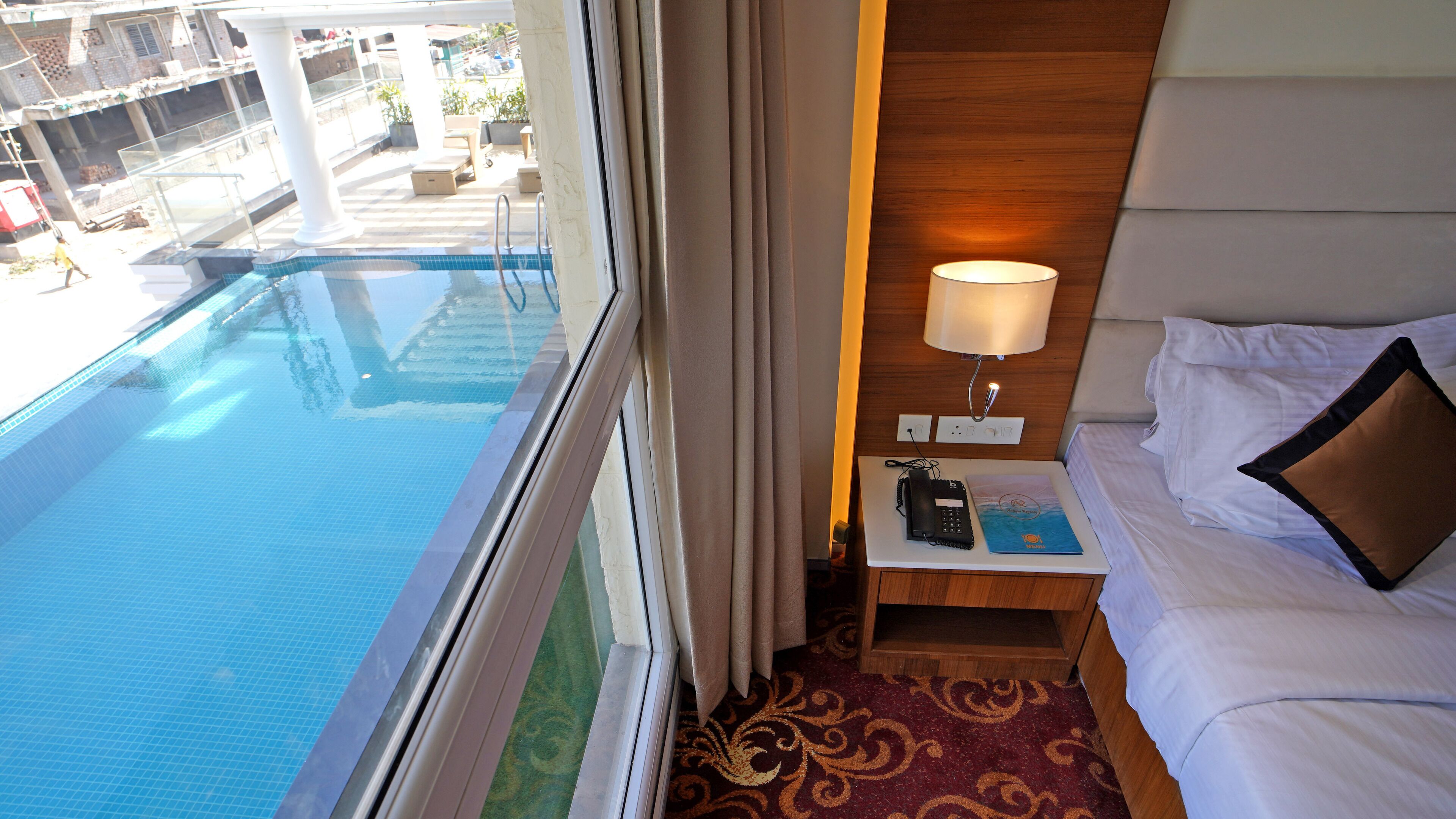 Habitación de hotel con vista a una piscina, una mesita de noche con lámpara, teléfono y un libro.