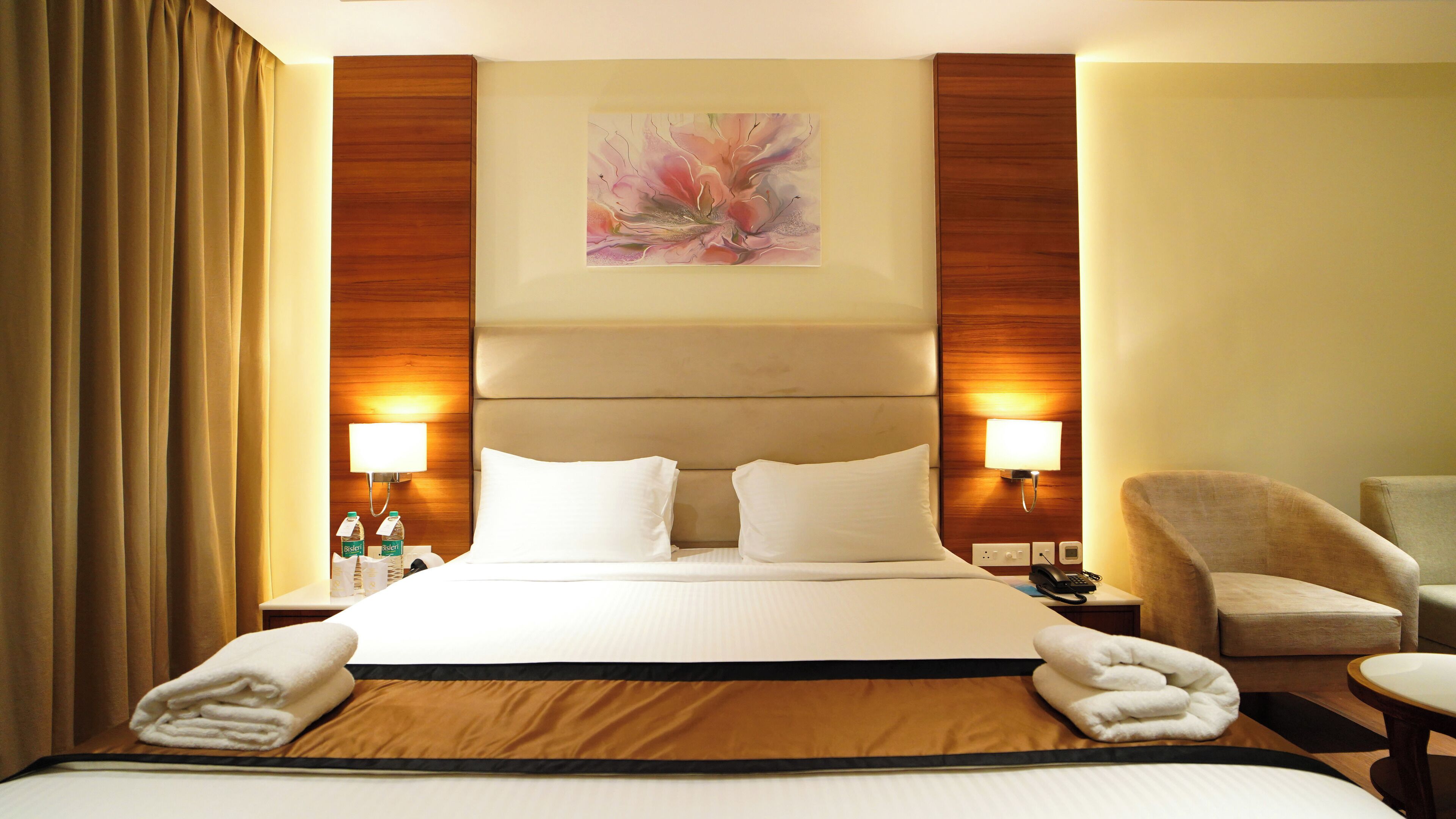 A hotel room with a large bed, two bedside lamps, a painting above the bed, and a chair to the right.