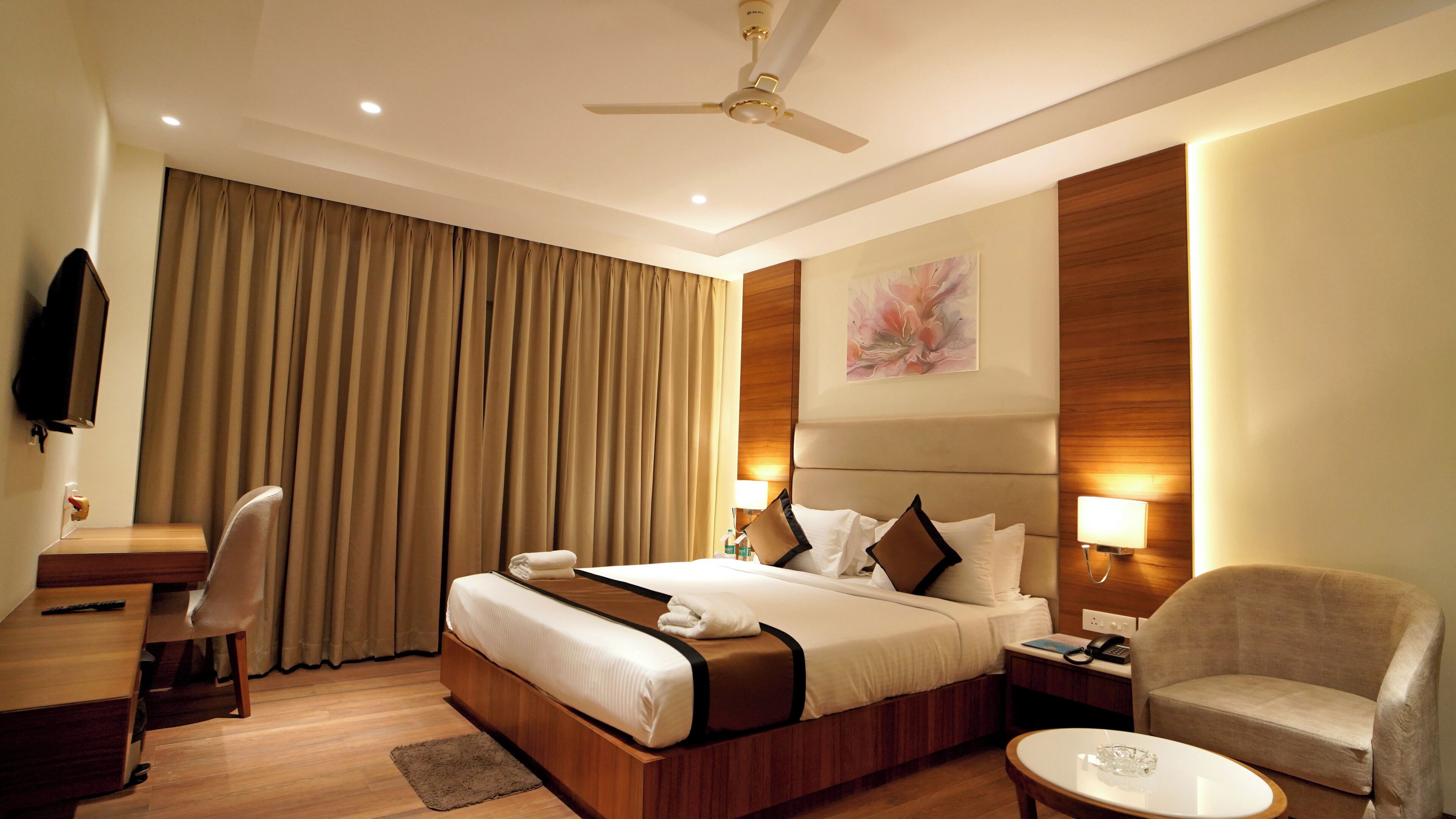 Quarto de hotel com cama, escrivaninha, cadeira, mesa pequena e ventoinha no teto.