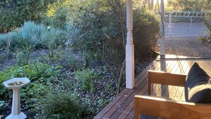 Una terrassa de fusta amb una banca i una banyera per a ocells, envoltada de vegetació abundant i una tanca metàl·lica.