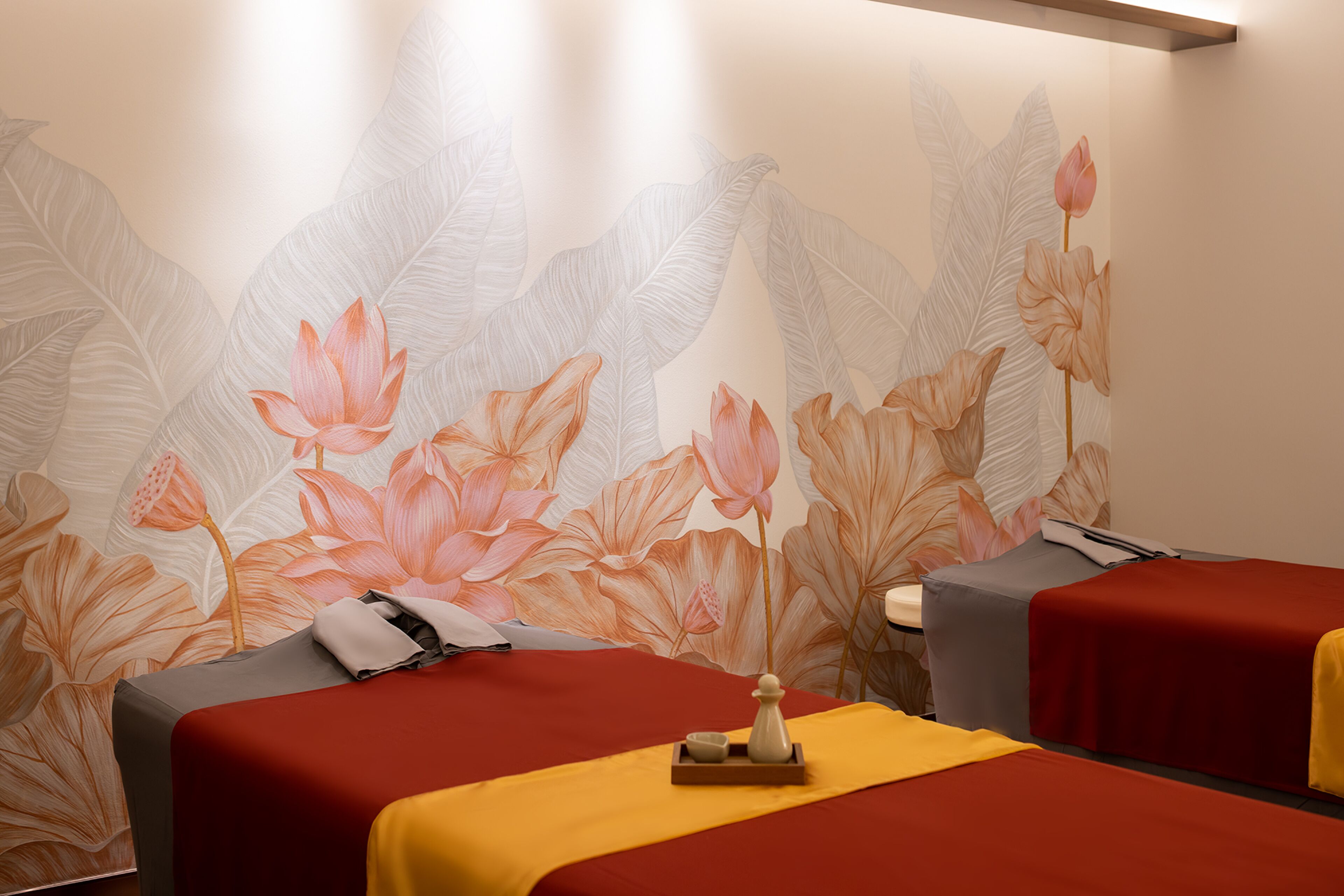A room with two beds, each with a red bedspread and a yellow blanket. There is a small table with a vase and a candle between the beds. The wall behind the beds features a large floral wallpaper.
