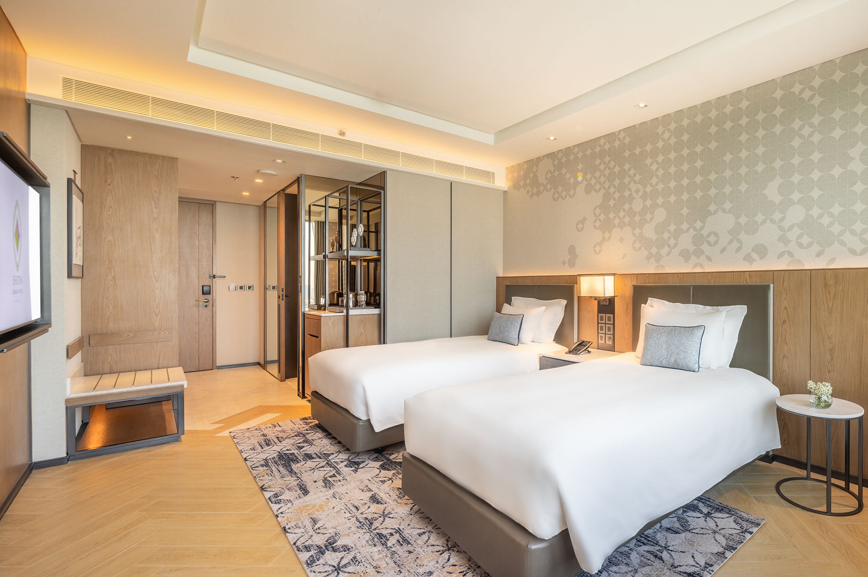 A modern hotel room with two beds, a small seating area, a TV, and a bathroom with a mirror and vanity.