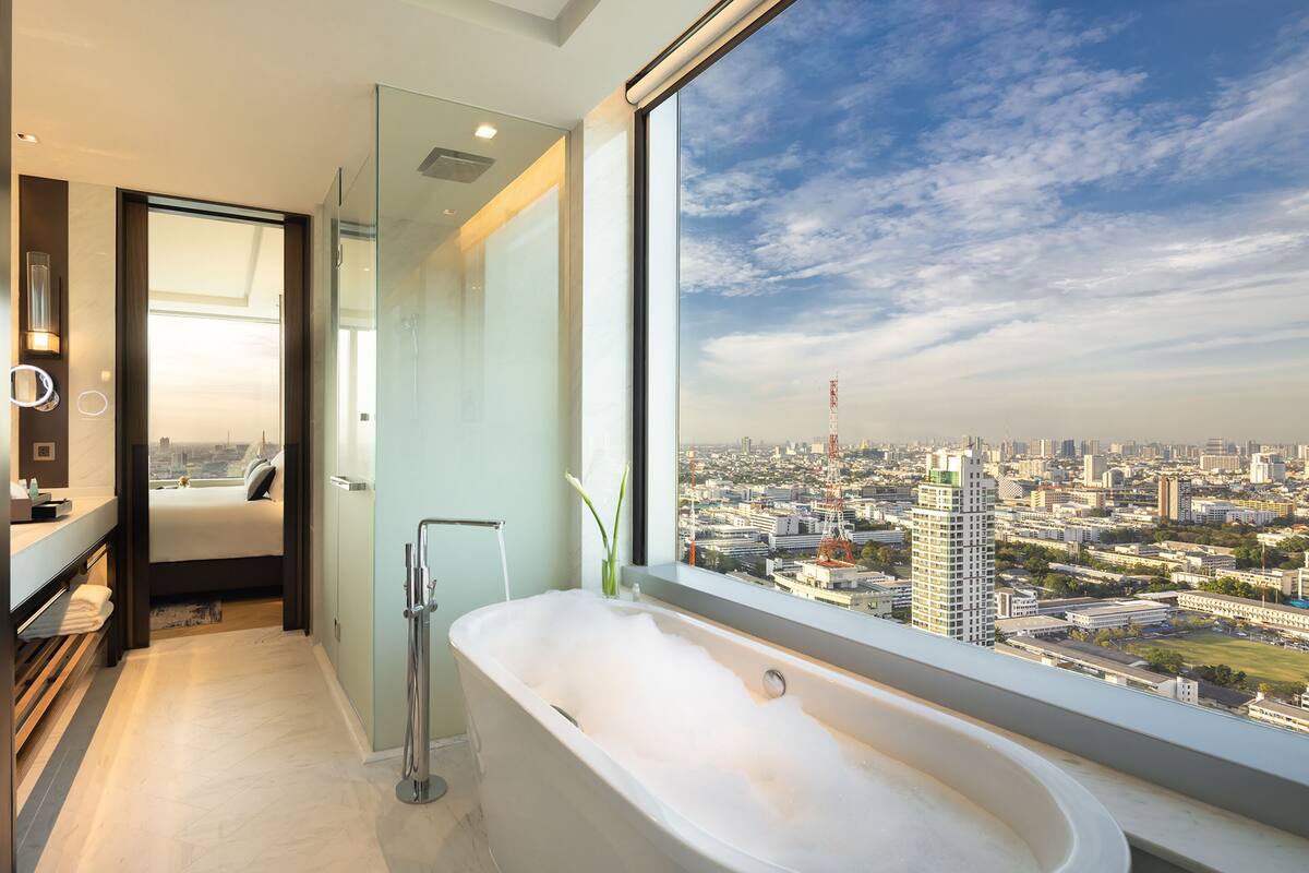 A modern bathroom with a large bathtub, a glass shower enclosure, and a city view through a large window.