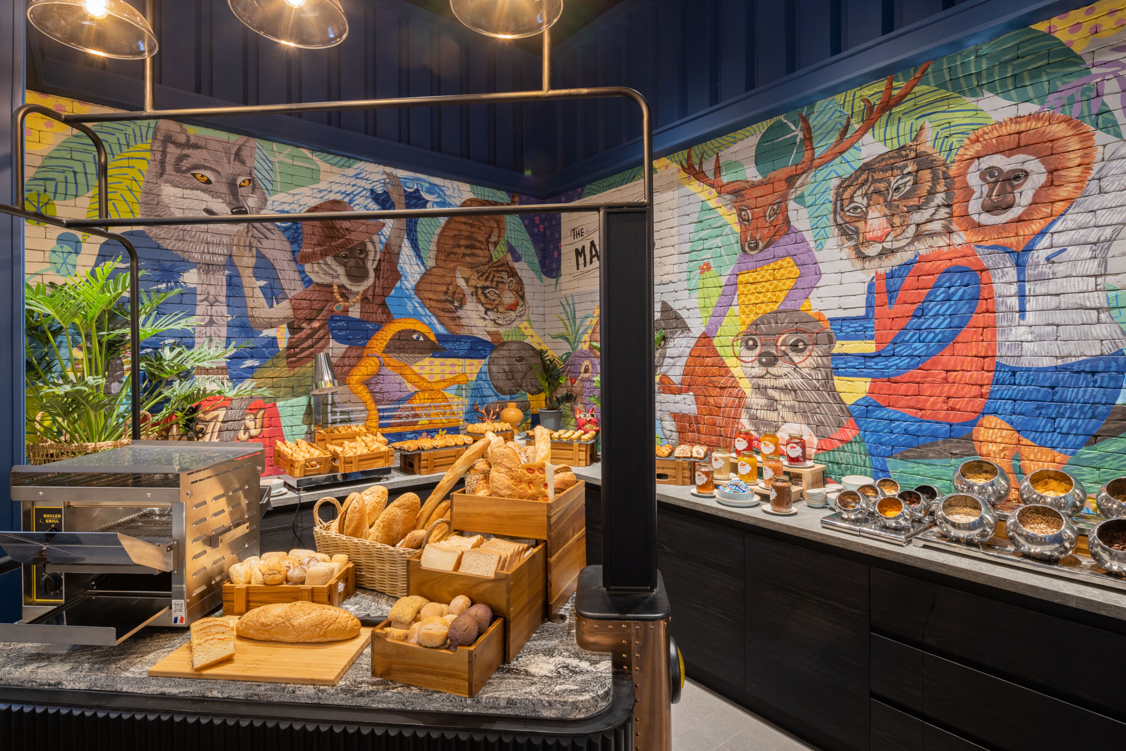 Mostrador de panadería con una variedad de panes y pasteles, una máquina de café y un mural colorido con animales en la pared.