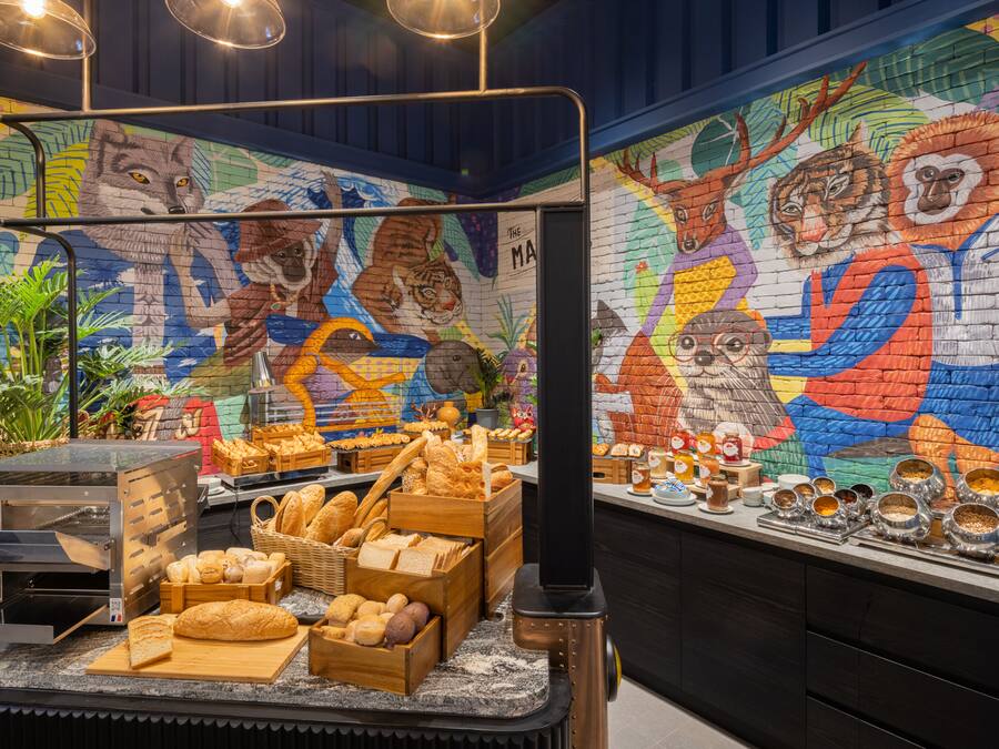 Mostrador de panadería con una variedad de panes y pasteles, una máquina de café y un mural colorido con animales en la pared.