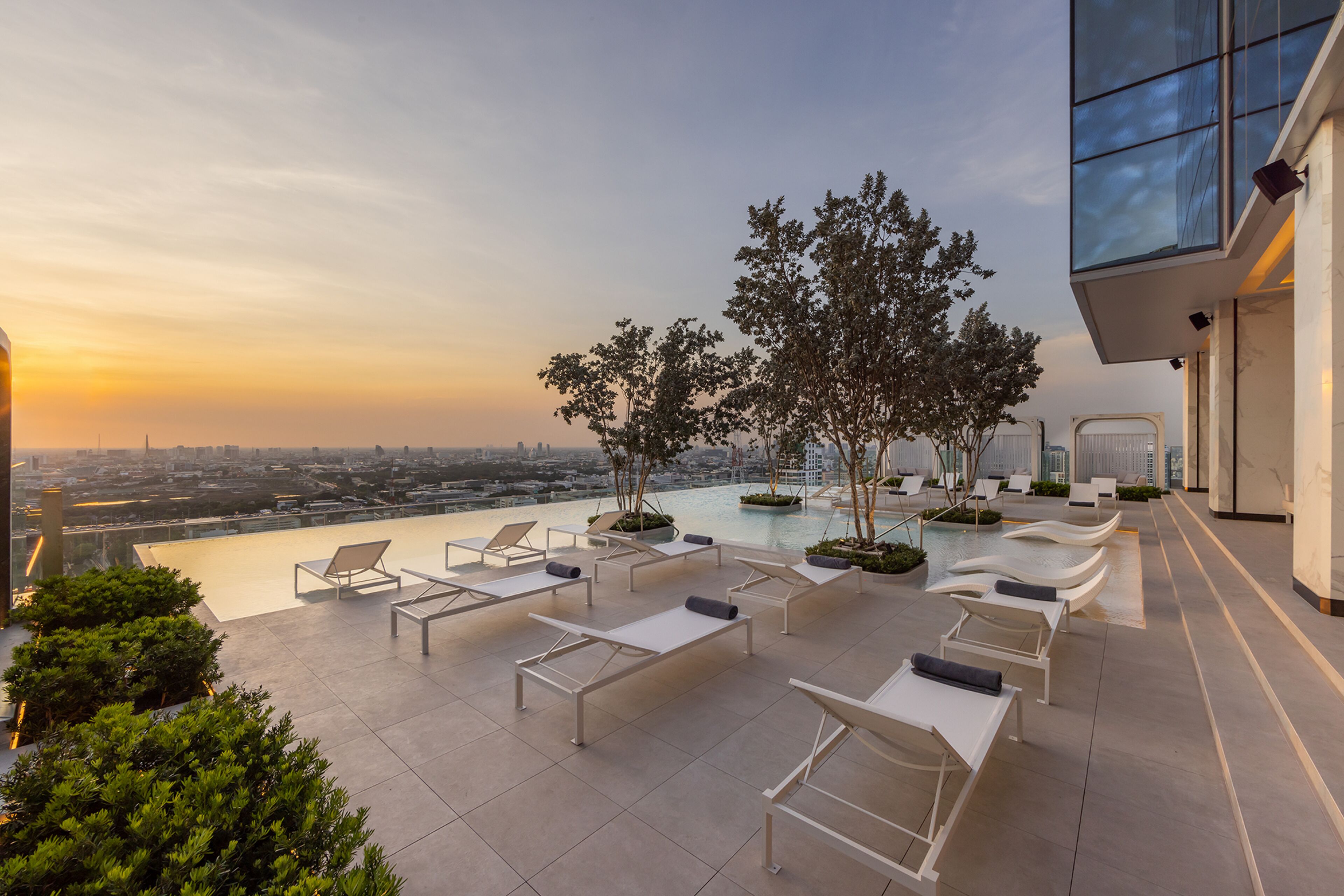 A rooftop lounge with lounge chairs and a swimming pool, overlooking a cityscape at sunset.