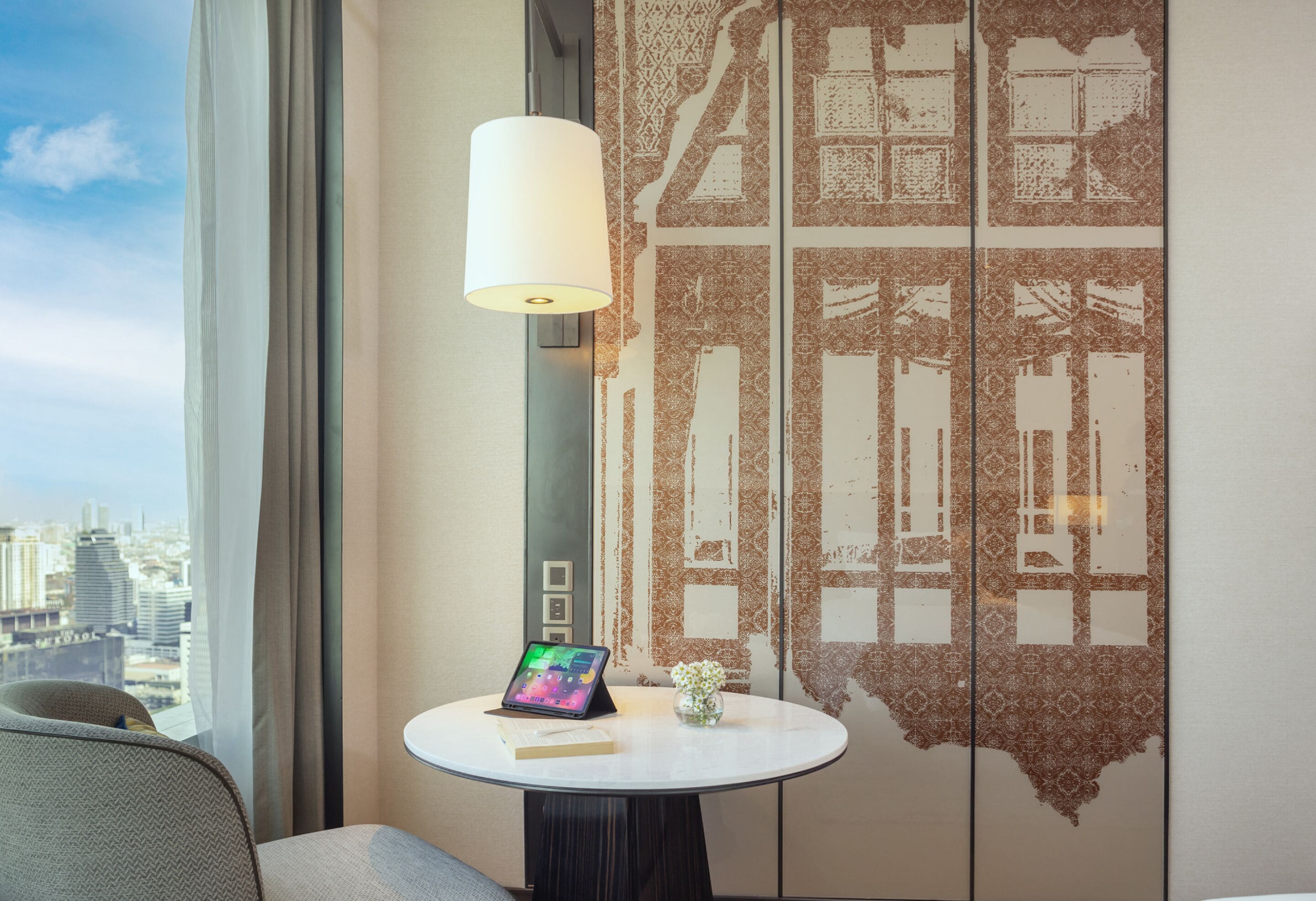 A hotel room with a view of the city, a round table with a lamp, a laptop, and a small vase with flowers.