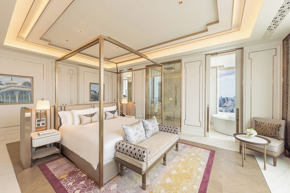 A luxurious bedroom with a four-poster bed, a sitting area with a sofa, a small table, and a view of the city through a glass door.
