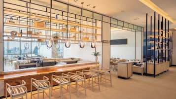 A modern bar area with a wooden counter, metal chairs, and a city view through large windows.