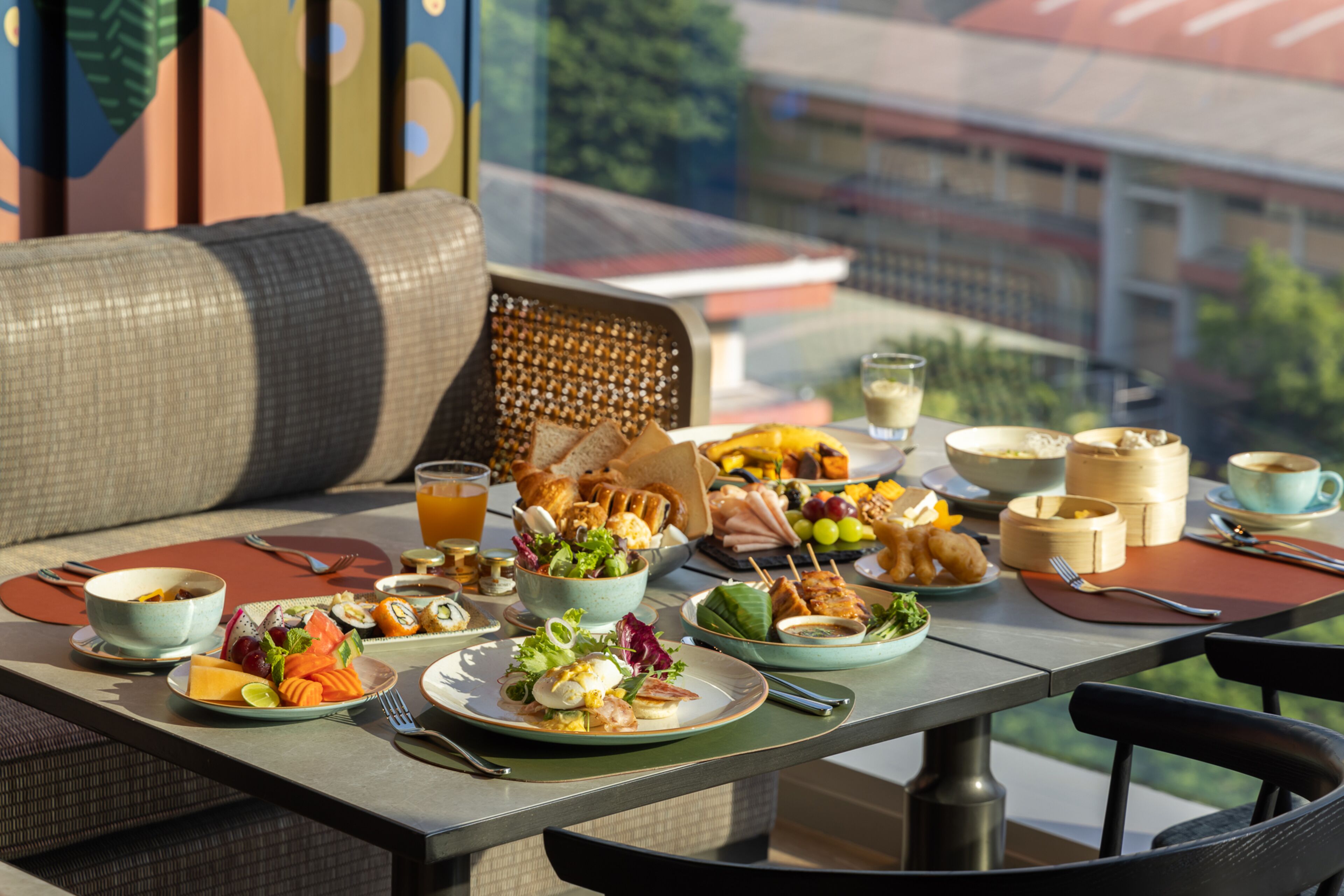 A dining table with a variety of dishes, including salads, fruits, and bread, set on an outdoor patio with a view of buildings in the background.