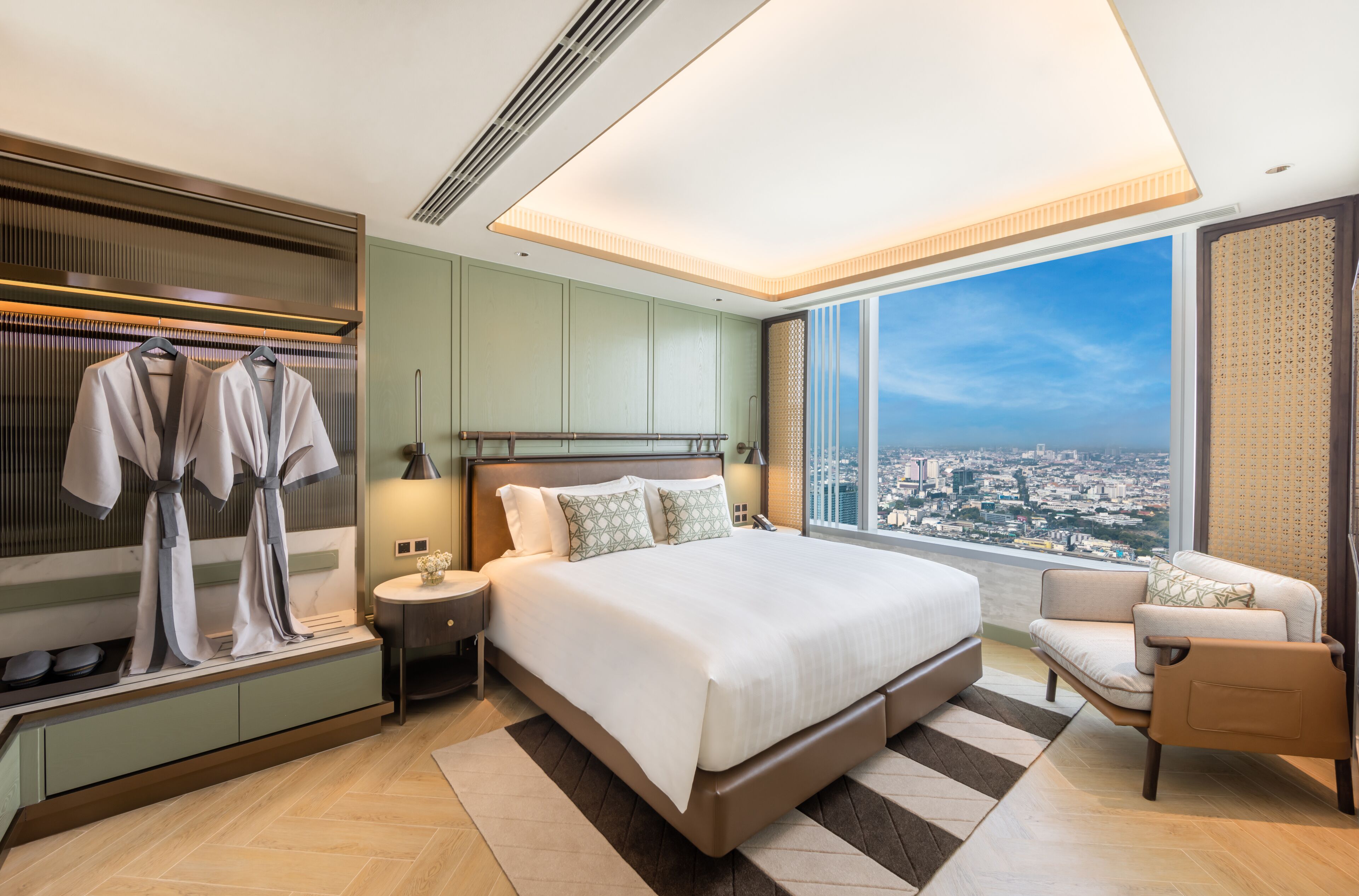 A modern hotel room with a large bed, a view of the city, a seating area, and a wardrobe with hanging clothes.