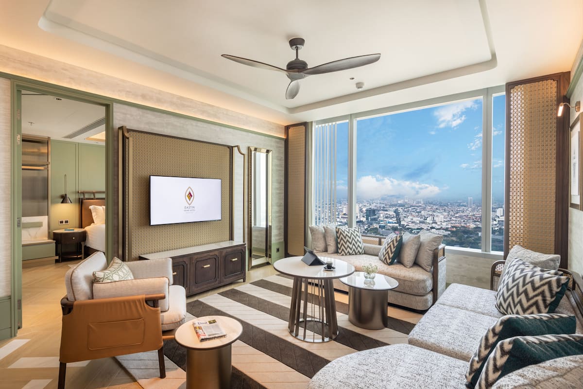 A modern hotel room with a large city view, a flat-screen TV, and a variety of seating options including a sofa, armchair, and a small round table.